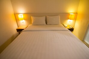 A neatly made bed with two pillows, flanked by two bedside tables with lamps, in a room with plain walls and a window.