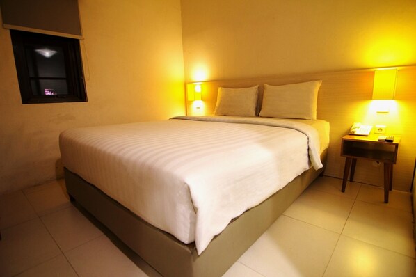 A neatly made bed in a hotel room with a small bedside table, a lamp, and a window with curtains. - Fovere Puri Semarang (Semarang)