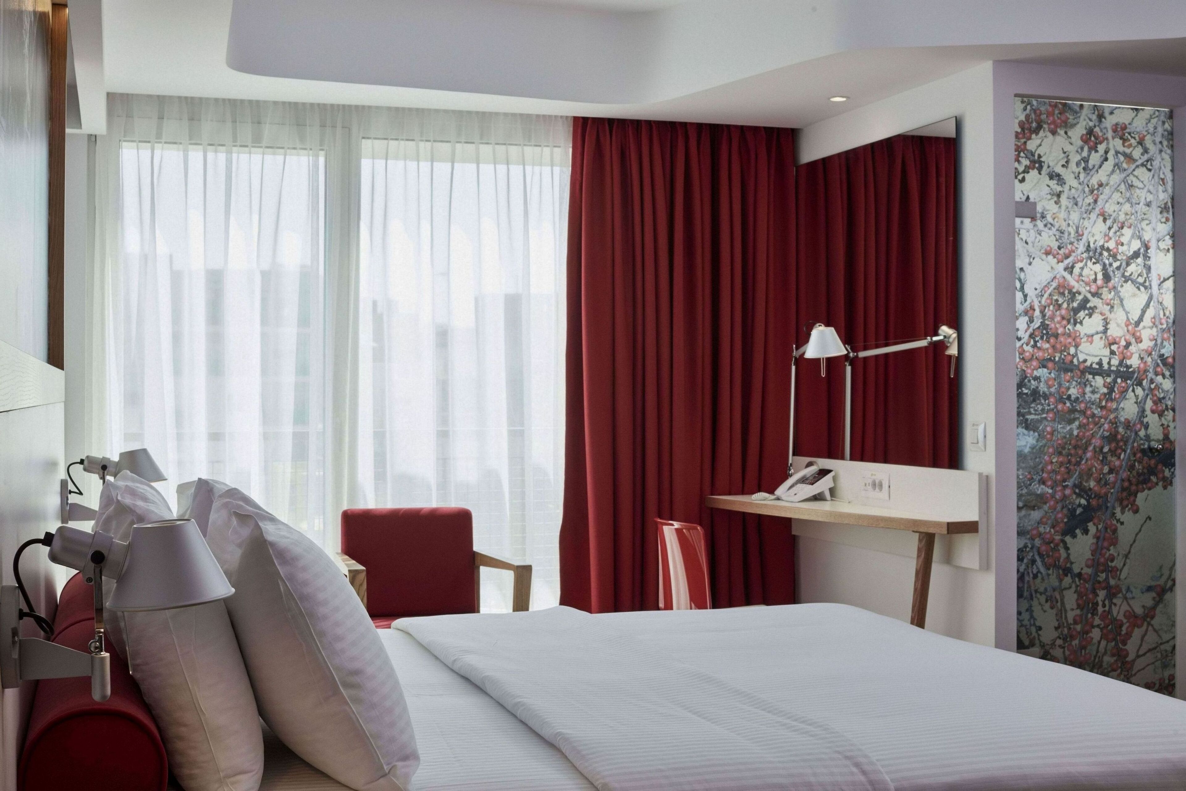 Een hotelkamer met een bed, een bureau, een stoel, een lamp en een groot raam met gordijnen.