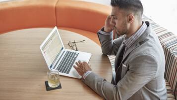 A person sitting at a round wooden table with a laptop, a glass of drink, and a pair of glasses on the table.