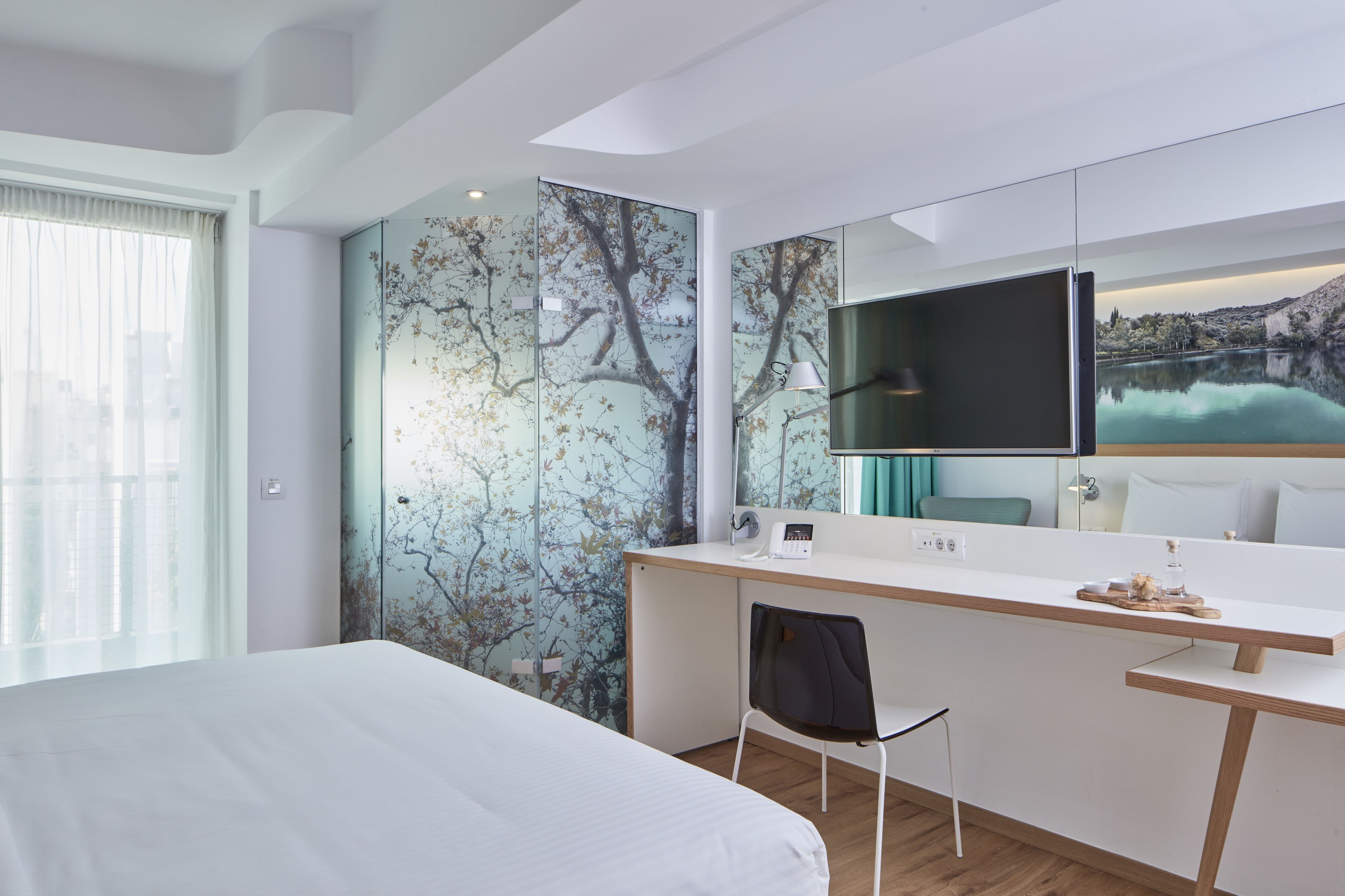 A modern hotel room with a large bed, a desk with a chair, a flat-screen TV, and a large window with a view of a lake and mountains.