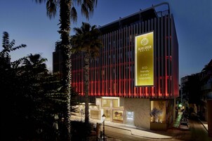 Hotel verde-olive com fachada moderna, sinalização destacada e entrada bem iluminada.