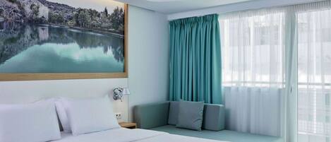 A hotel room with a bed, a seating area with cushions, a large window with curtains, and a painting of a landscape on the wall.