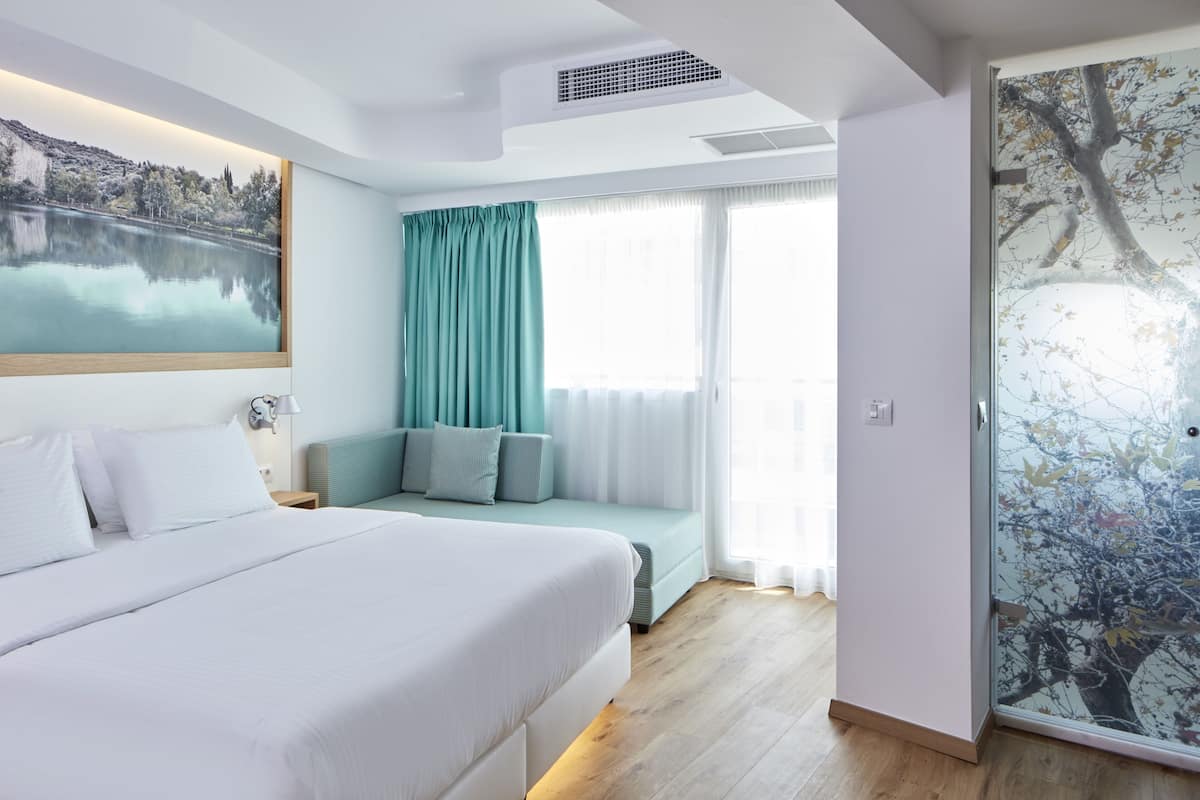 Une chambre d’hôtel moderne dotée d’un grand lit, d’un coin salon avec vue sur un lac et d’une porte en verre ornée d’une fresque représentant un arbre.