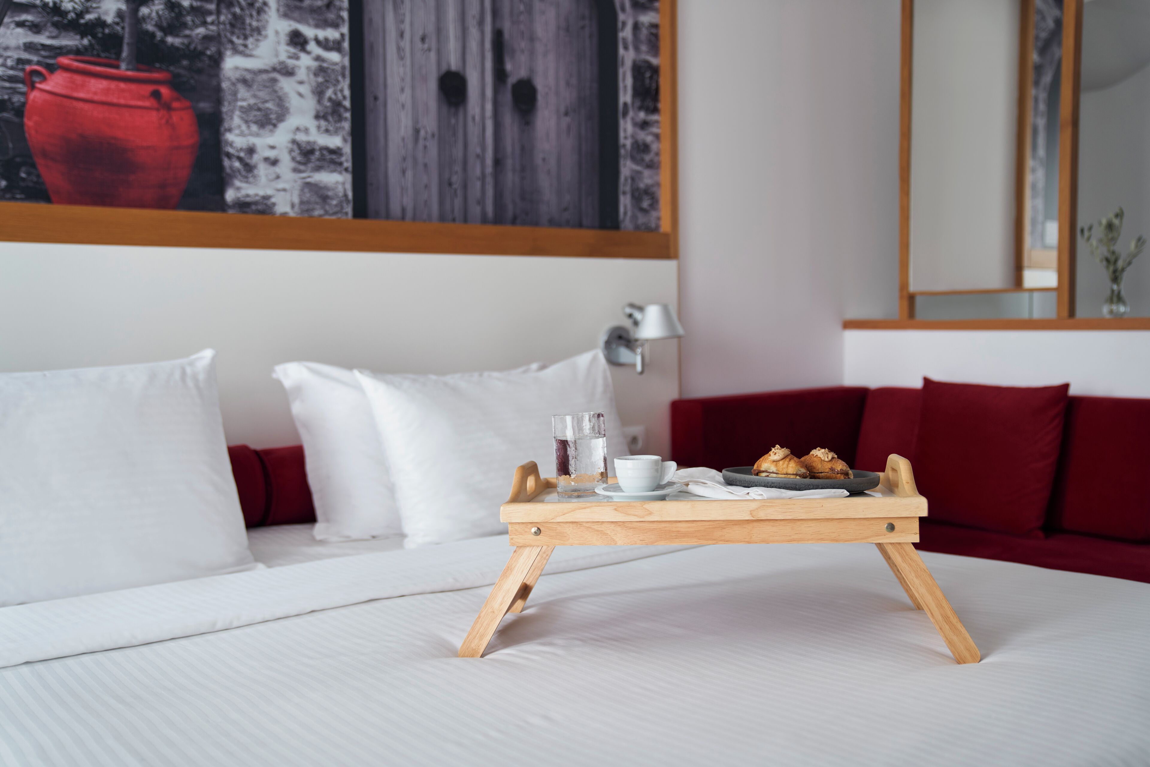 A neatly made bed with white linens, a wooden tray table with breakfast items, and a red sofa with white cushions.