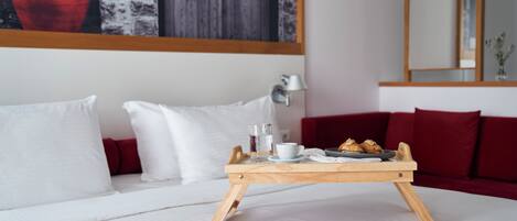A neatly made bed with white linens, a wooden tray table with breakfast items, and a red sofa with white cushions.