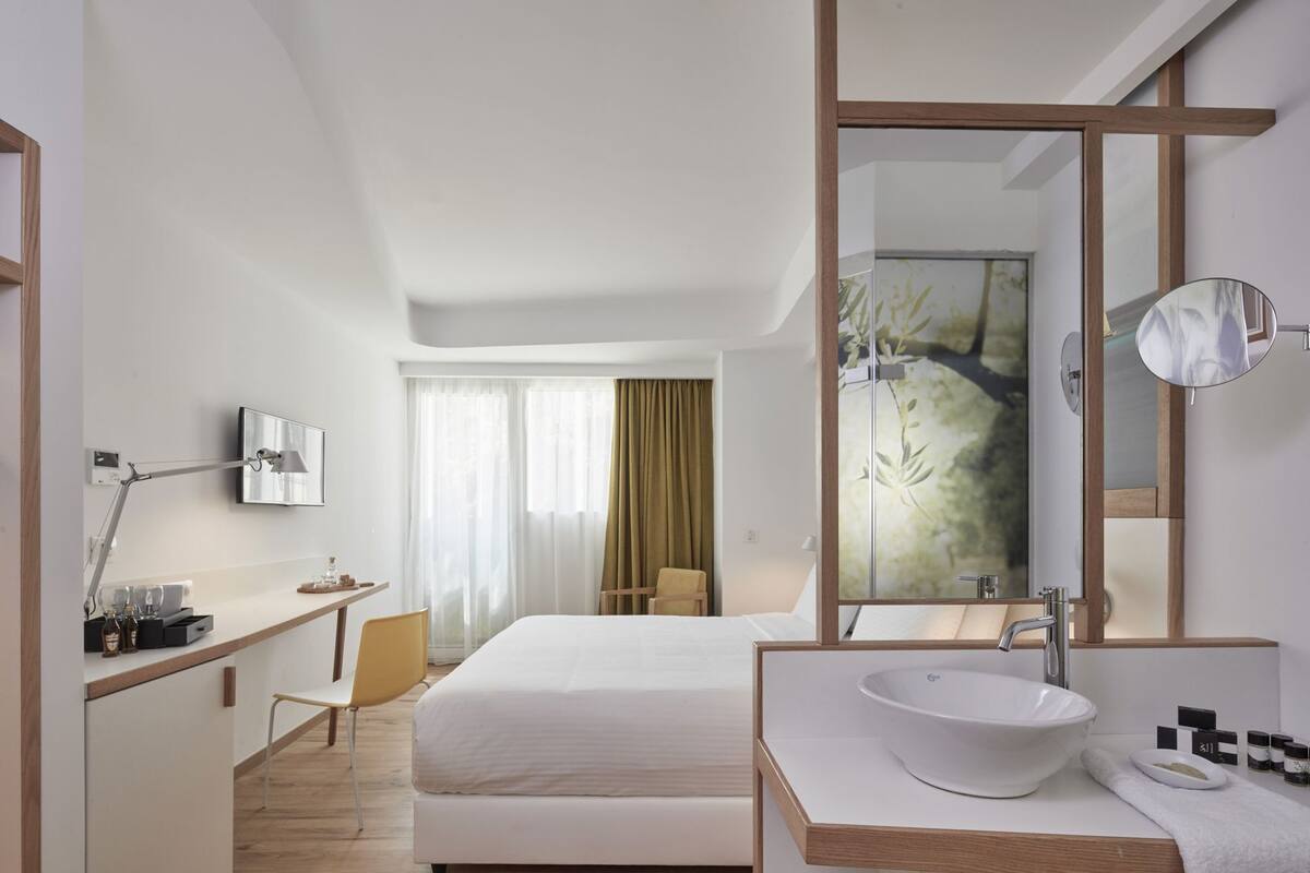 Une chambre d’hôtel comprenant un lit, un bureau avec une lampe, un miroir, une salle de bain avec un lavabo et une chaise.