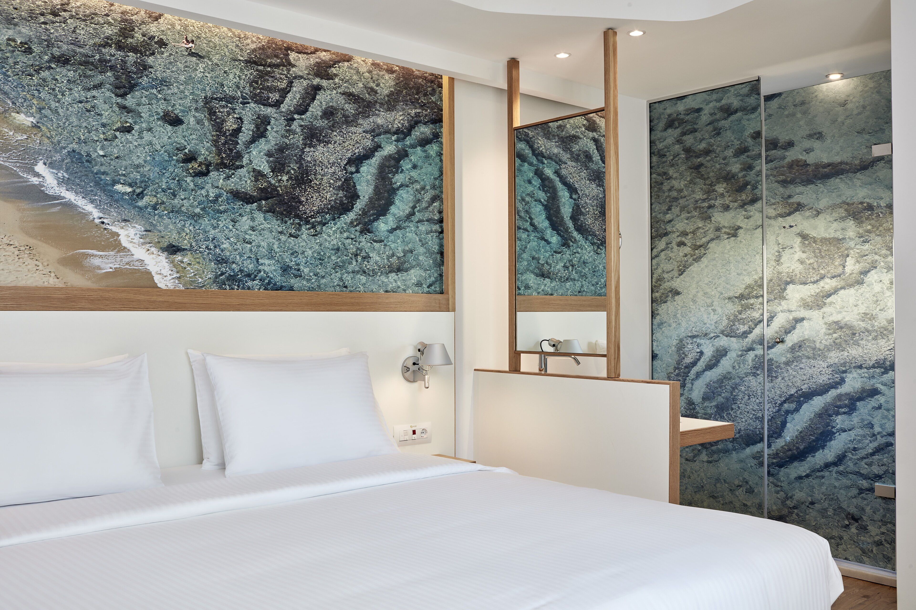 Ein modernes Hotelzimmer mit einem großen Bett, weißen Leinentextilien, einem wandmontierten Spiegel und einem strukturierten Wandbild.