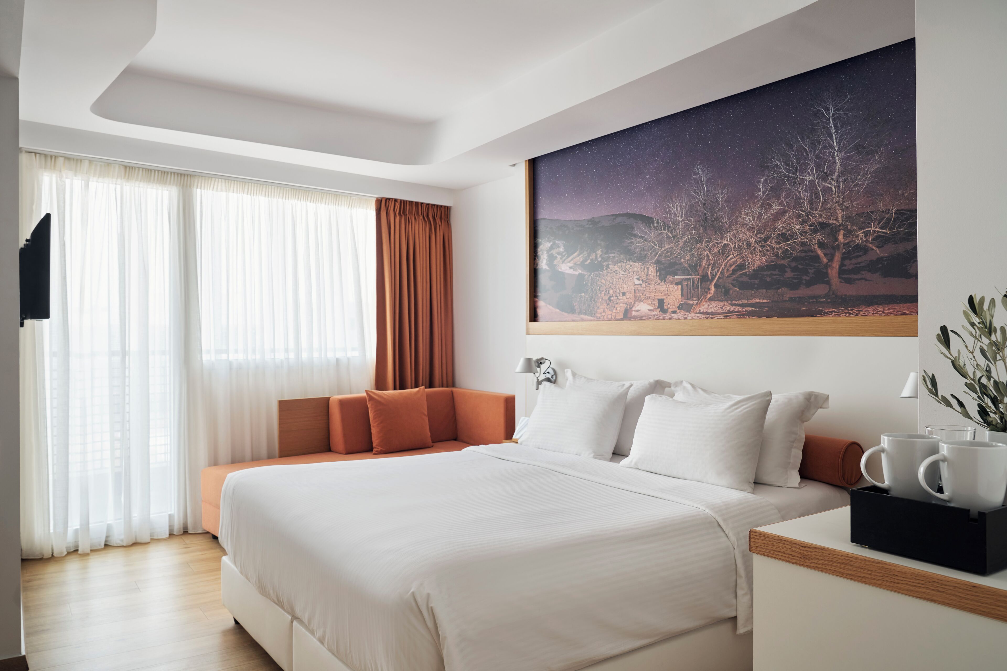 Una habitación de hotel moderna con una cama grande, un televisor de pantalla plana, un mural del cielo nocturno y una mesita de noche con un florero y dos tazas.