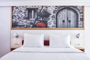 Quarto com cama, almofadas brancas, uma almofada decorativa vermelha, duas mesas de cabeceira com abajures e um quadro na parede retratando um edifício de pedra.