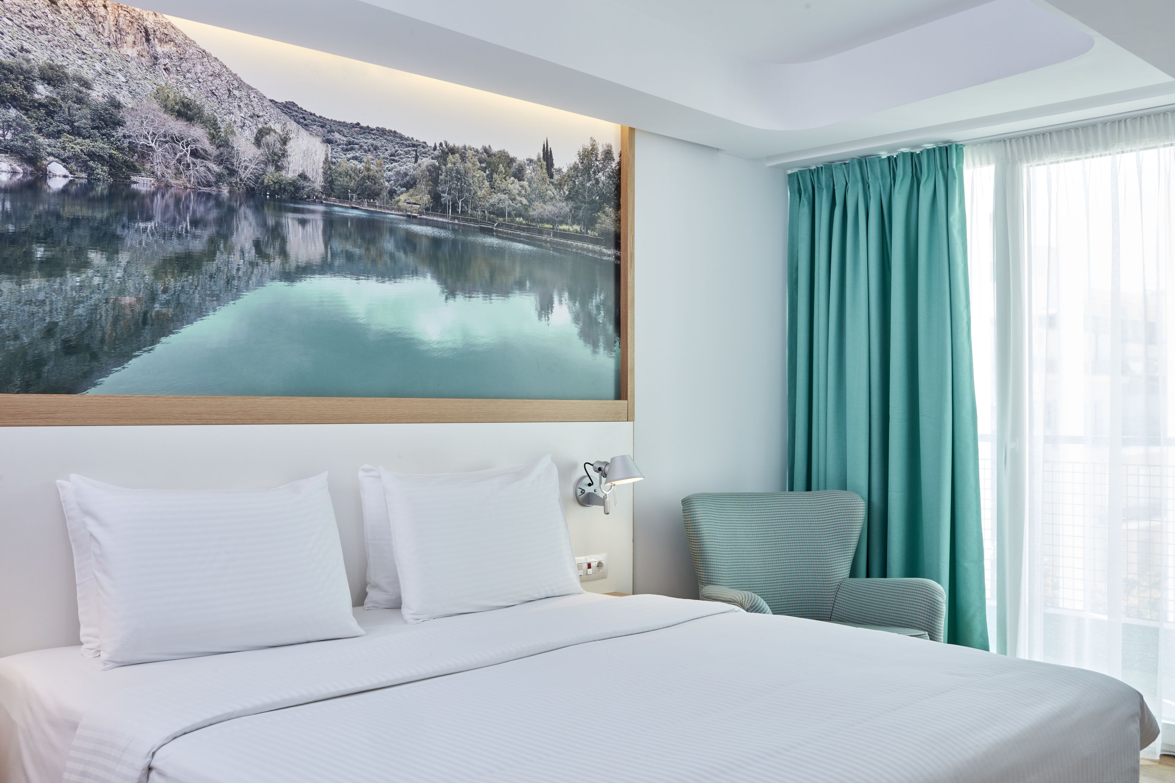 A hotel room with a large bed, a view of a lake, a window with curtains, and a wall-mounted lamp.