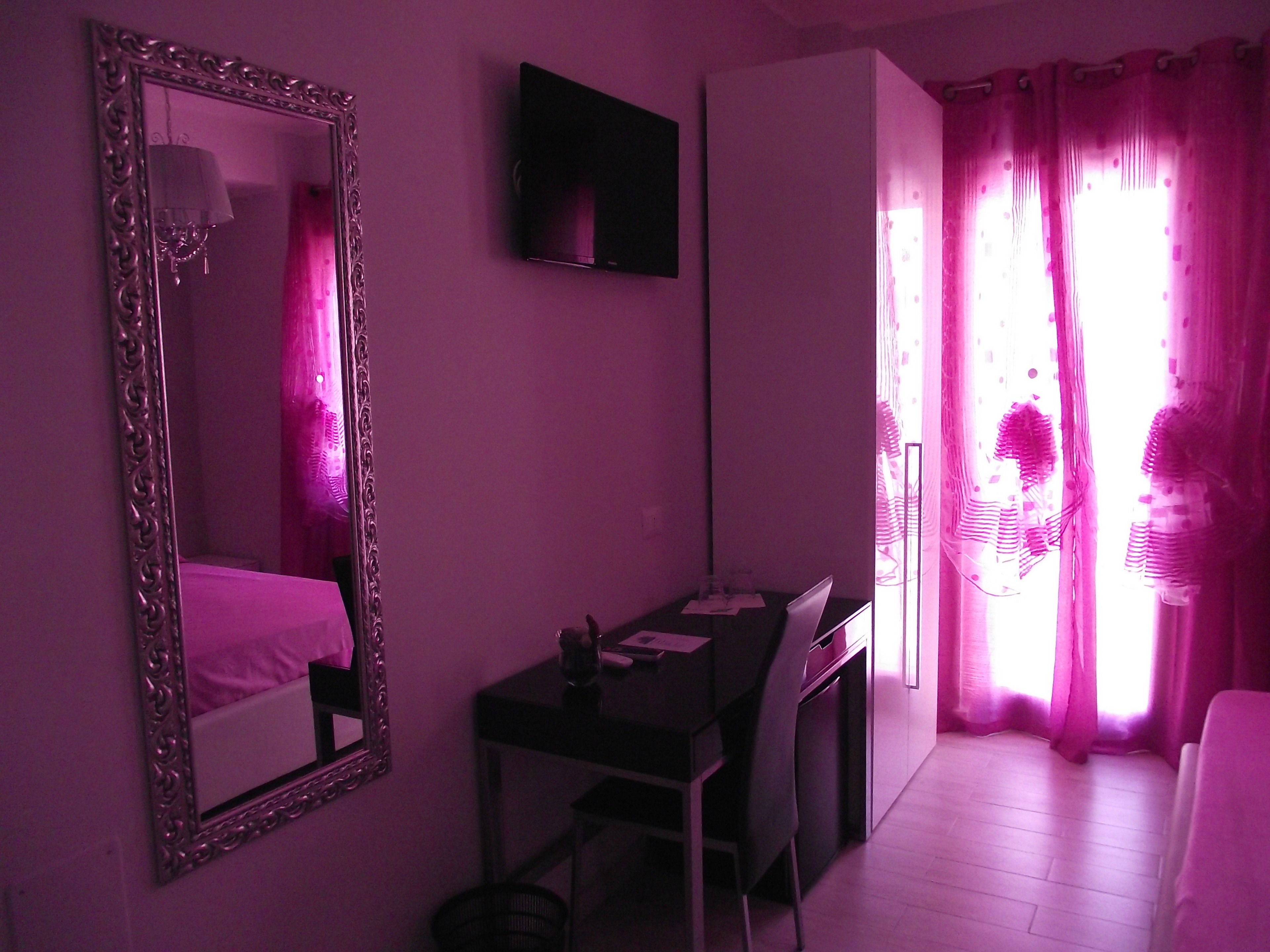 Une chambre dotée d’un grand miroir, d’un téléviseur à écran plat, d’un bureau avec une chaise et d’une fenêtre ornée de rideaux roses.