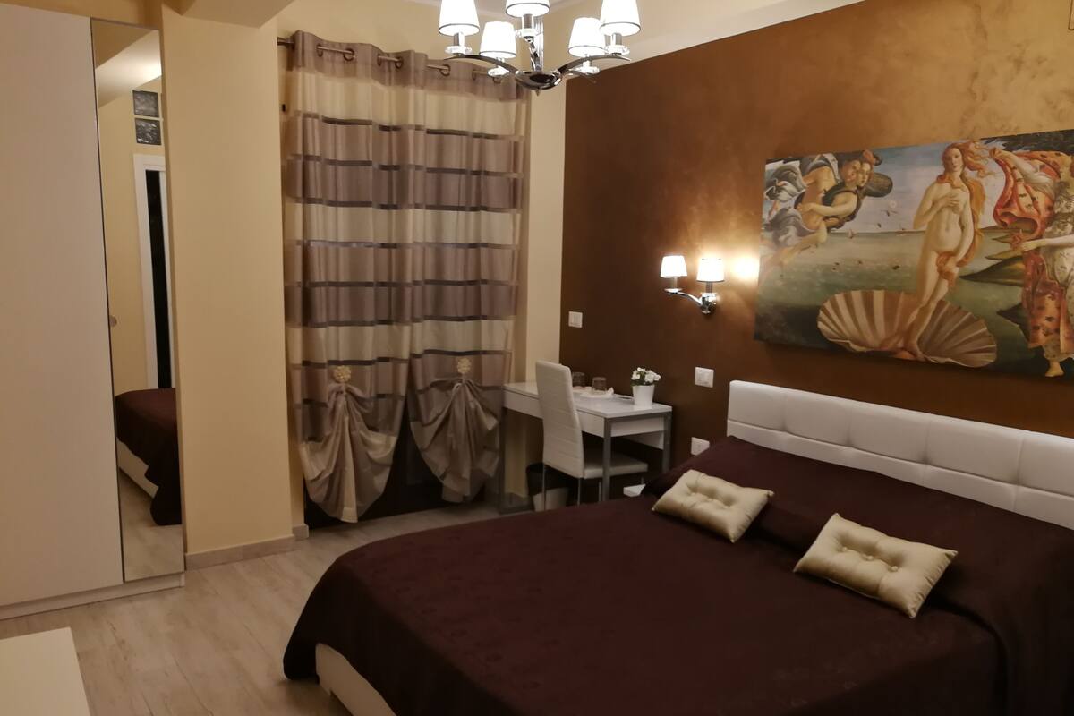 Une chambre d’hôtel avec un grand lit, un bureau et une chaise. Il y a un tableau au mur et un lustre.