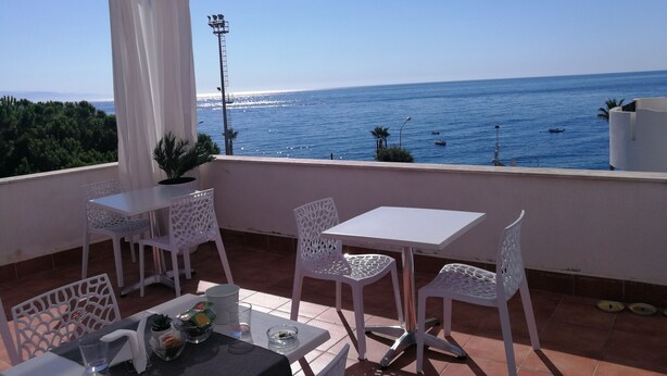 A terrace with white tables and chairs, a potted plant, and a view of the sea. - B&B Il Mare degli Dei (Letojanni)