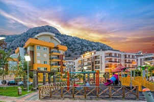 Un parque infantil con toboganes y estructuras para escalar, rodeado de edificios de varios pisos con balcones y un paisaje montañoso de fondo.