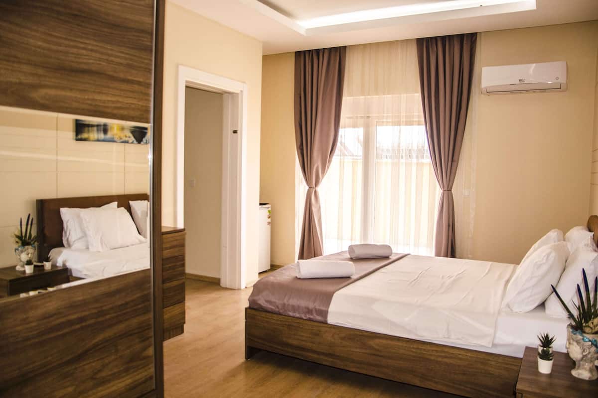 A hotel room with a wooden bed, a bedside table with a lamp, a small potted plant, and a window with curtains.
