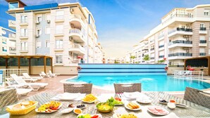 Un espace repas aménagé au bord de la piscine, avec une table dressée pour le petit-déjeuner, donnant sur une piscine, et des bâtiments modernes en arrière-plan.