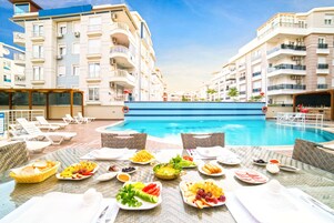 A poolside dining area with a table set for breakfast, overlooking a swimming pool, with modern buildings in the background.