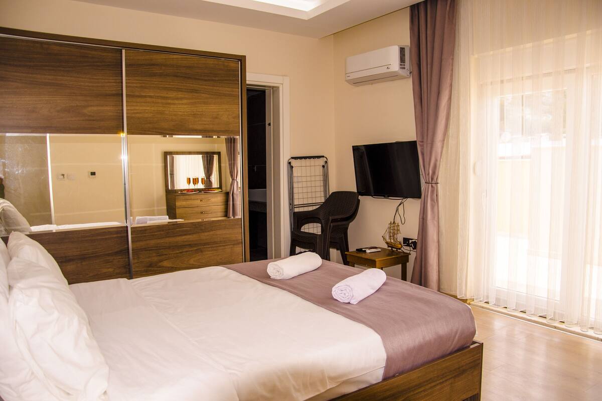 A hotel room with a bed, a mirror, a TV, a desk, and a window with curtains.