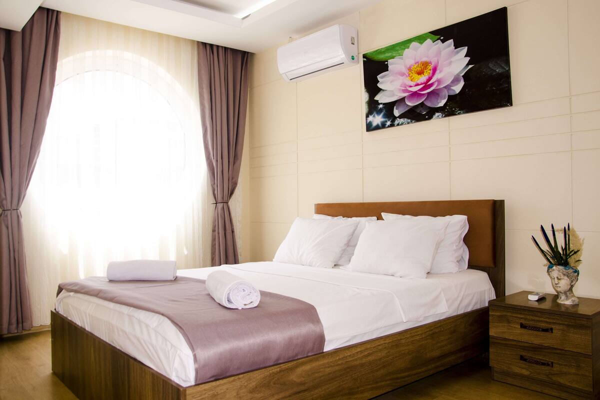 A neatly made bed with white linens and a purple throw, a wooden nightstand with a vase, and a wall-mounted air conditioner.
