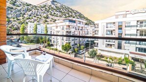 Un balcon avec des chaises et une table en plastique blanc, d’où l’on peut admirer un paysage urbain avec des immeubles et une montagne.