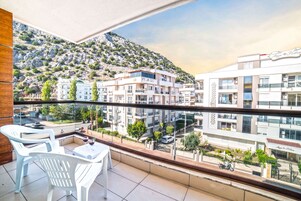 Un balcón con sillas y una mesa de plástico blanco, con vistas a un paisaje urbano que incluye edificios y una montaña.