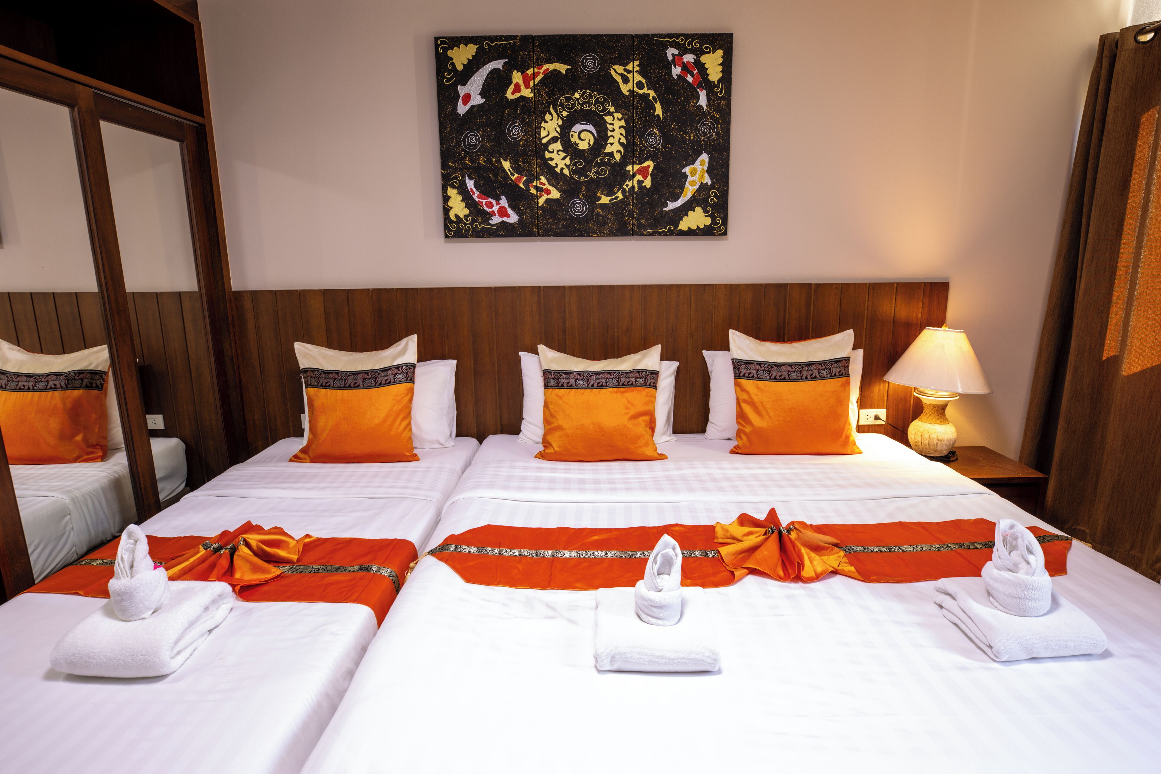 A hotel room with two beds, each with white linens and orange pillows. There is a painting on the wall above the beds, a nightstand with a lamp, and a wooden headboard.