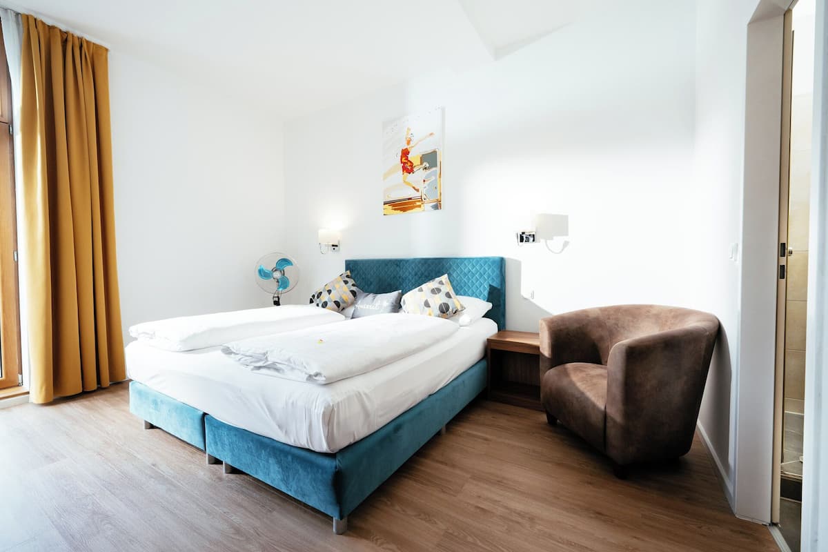 A modern hotel room with a large bed, a blue headboard, a brown armchair, and a wall-mounted fan.