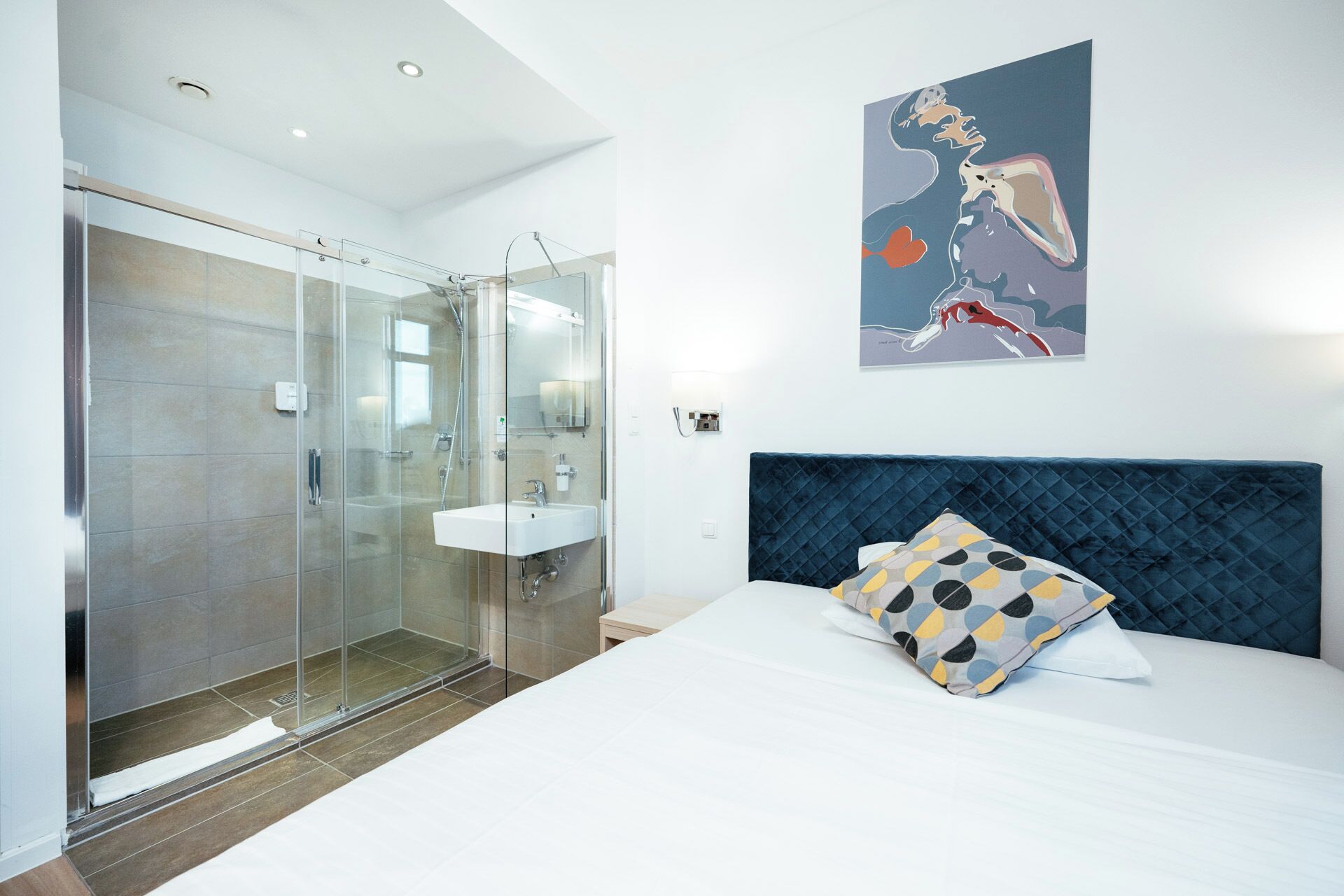 Ein modernes Hotelzimmer mit einer in Glas eingefassten Dusche, einem weißen Waschbecken und einem Bett mit dunkelblauem Kopfteil und einem Kissen mit Punkten.