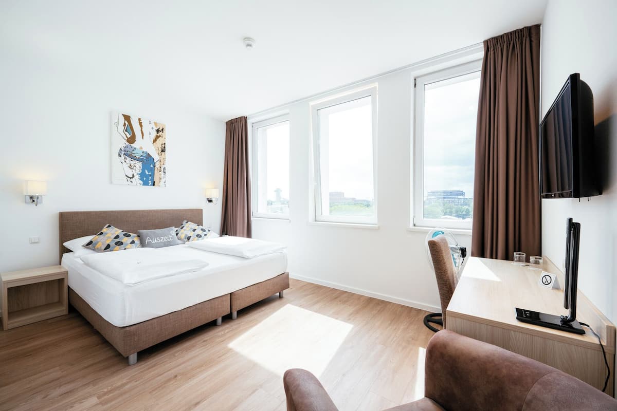 A modern hotel room with a bed, a desk, a chair, and large windows.