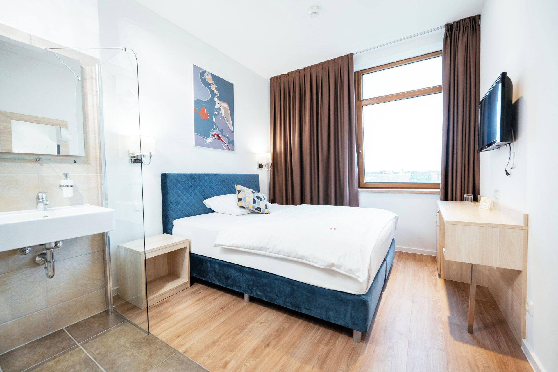 Una habitación de hotel moderna con una cama grande, una mesita de noche, un escritorio, un televisor y un baño con lavamanos y espejo.