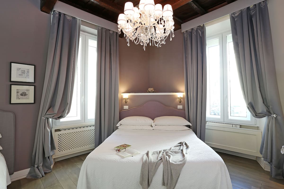 Ein ordentlich bezogenes Bett mit Buch und Brille, eine Chandelüster und ein Fenster mit Vorhängen.