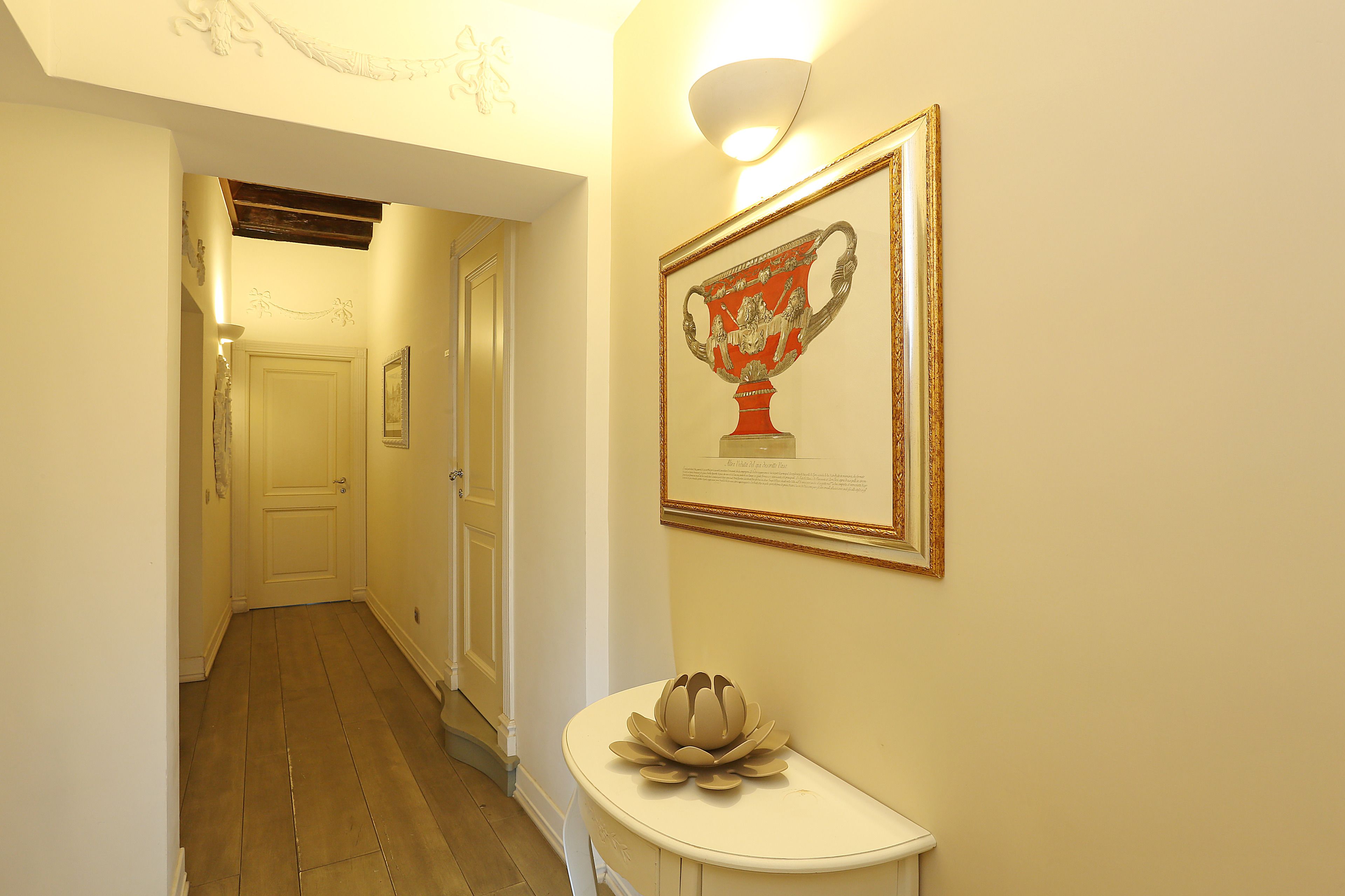 Un couloir avec une porte blanche au fond, un tableau encadré au mur et une console blanche sur laquelle est posé un objet décoratif.