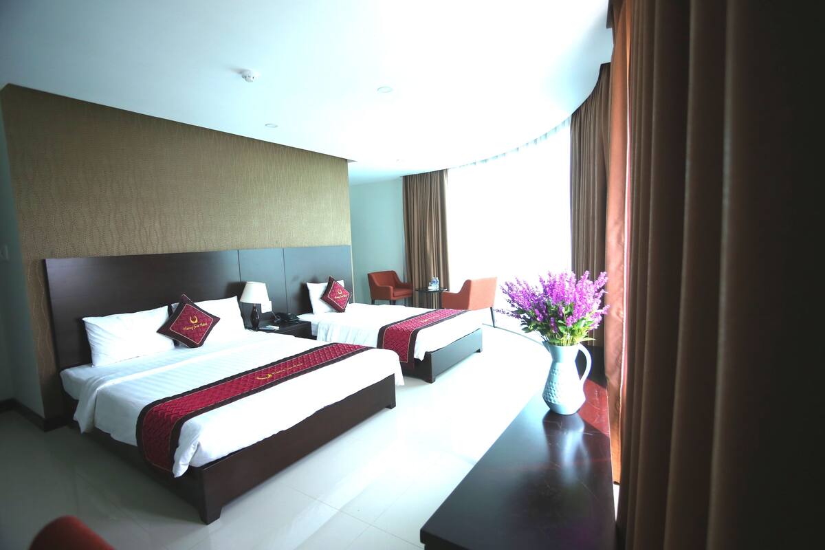 A hotel room with two beds, a large headboard, a desk, a chair, a vase with flowers, and a window with curtains.