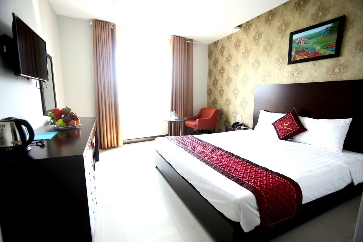 A hotel room with a large bed, a desk with a kettle, a chair, and a television.