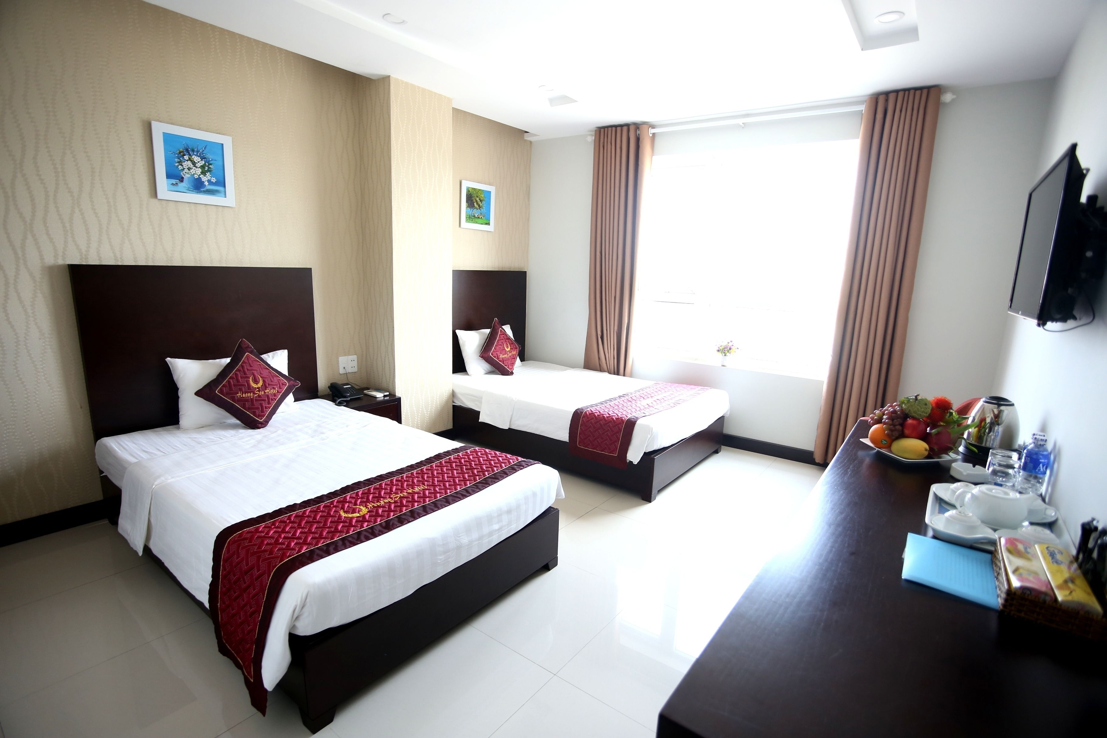 A hotel room with two beds, a desk with a fruit bowl, and a TV on the wall.