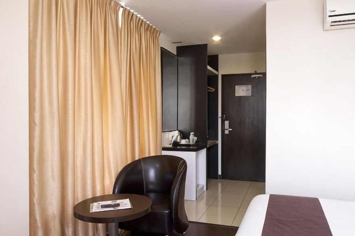 A hotel room with a bed, a small table with a chair, a desk with a chair, and a kitchen area in the background.
