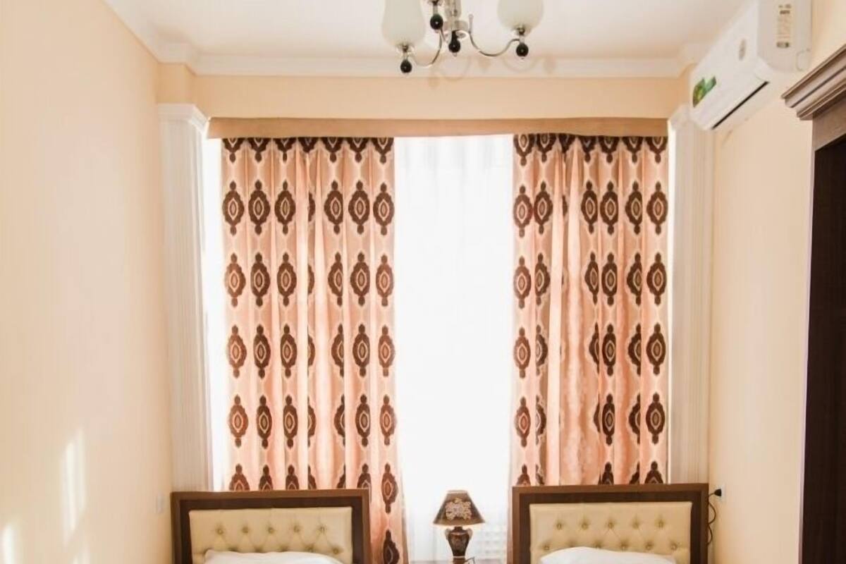A hotel room with two single beds, a patterned carpet, a chandelier, and a window with patterned curtains.