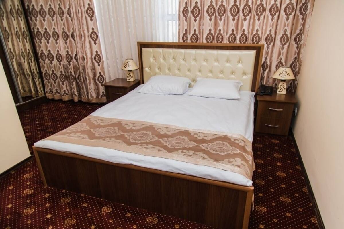A neatly made bed with a wooden frame and a tufted headboard, accompanied by two bedside tables with lamps, in a room with patterned curtains and a carpeted floor.