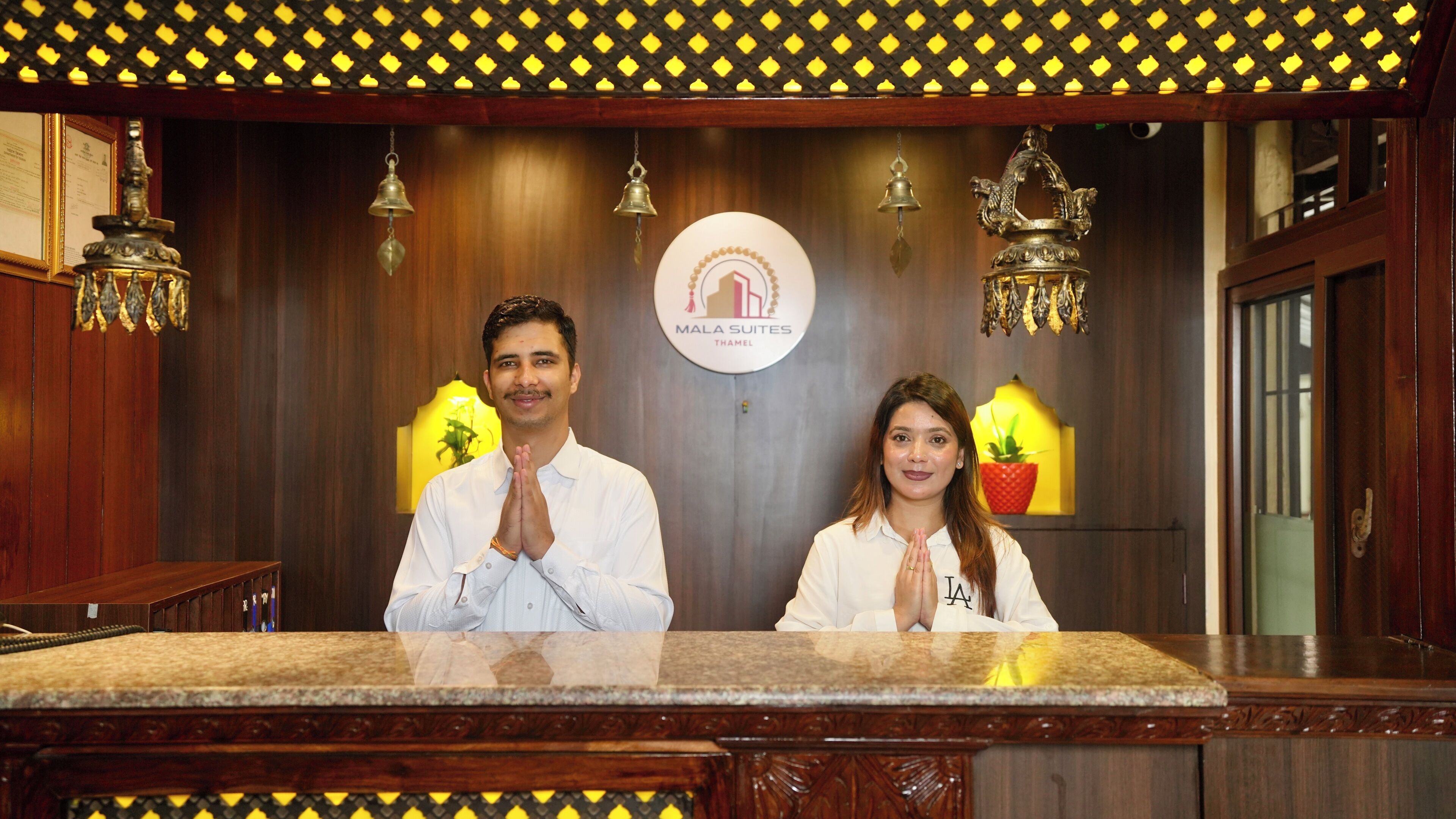A reception desk with a logo for 'Mala Suites Thamel' and decorative elements like hanging bells and a plant.