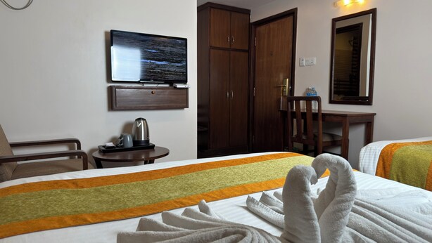 Quarto de hotel com cama, televisão fixada na parede, guarda-roupa de madeira, mesa pequena com chaleira e uma cadeira.