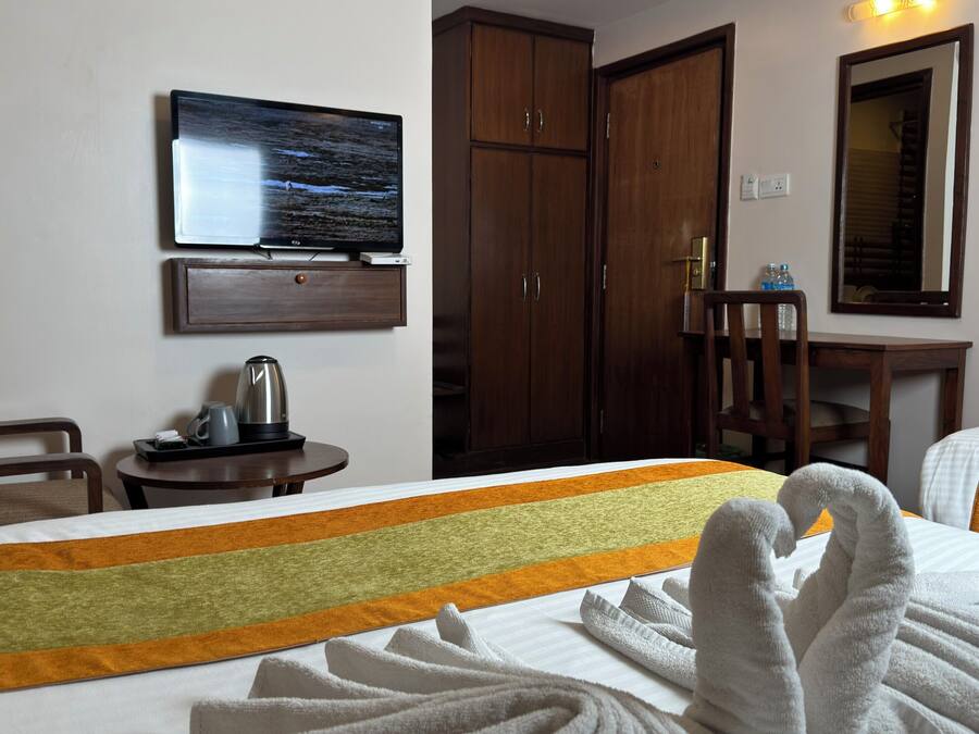 Habitación de hotel con cama, televisor montado en la pared, armario de madera, mesita con tetera y una silla.