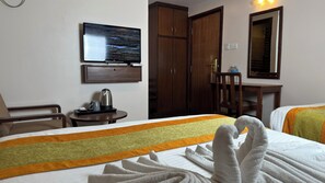 Habitación de hotel con cama, televisor montado en la pared, armario de madera, mesita con tetera y una silla.