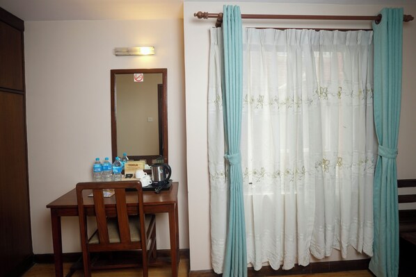 Una habitación con un escritorio de madera, una silla, un espejo, una botella de agua, un hervidor y una cortina con motivos florales.