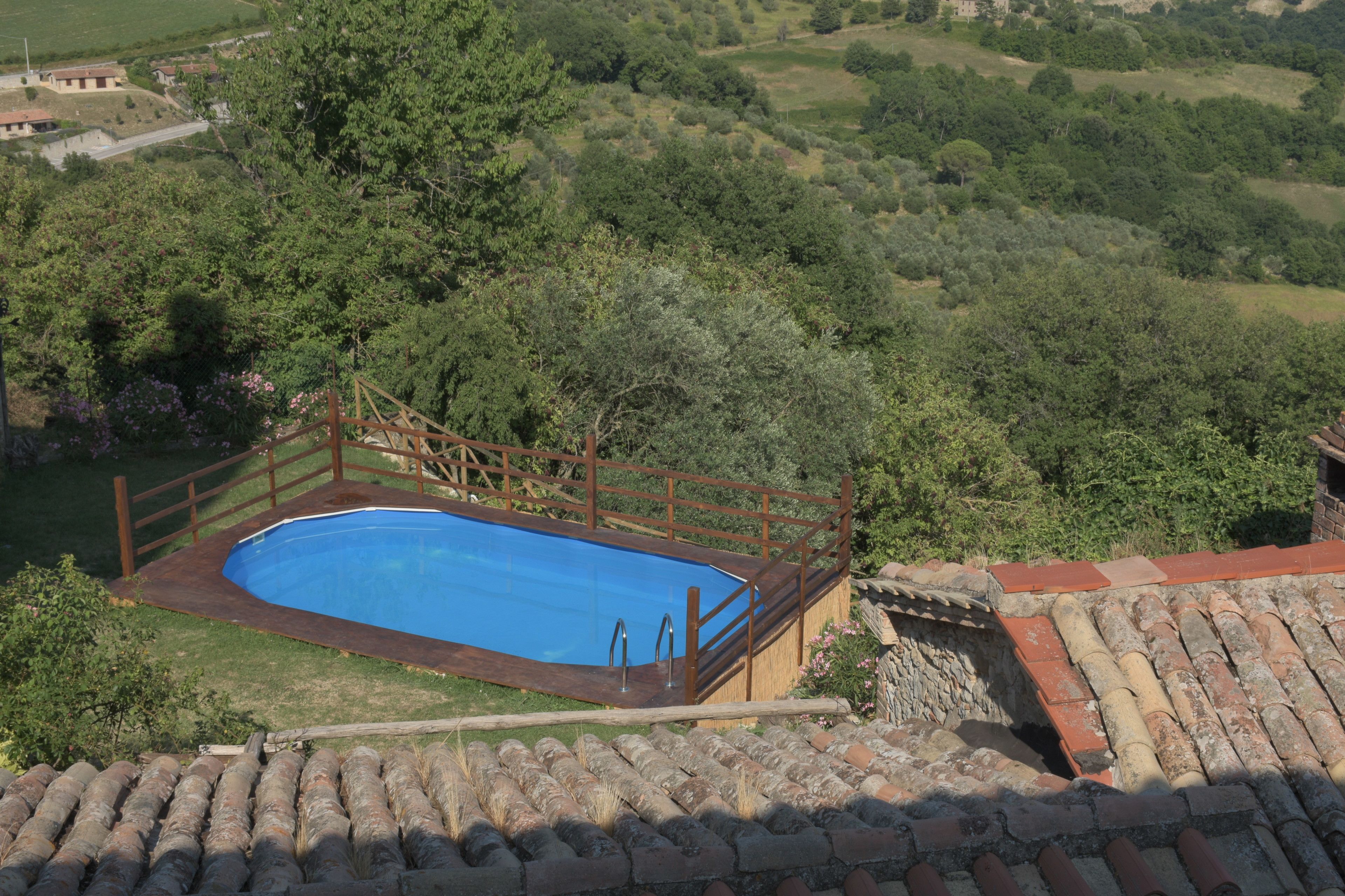 A swimming pool surrounded by a wooden deck and a stone wall, set in a lush green landscape with trees and hills in the background.