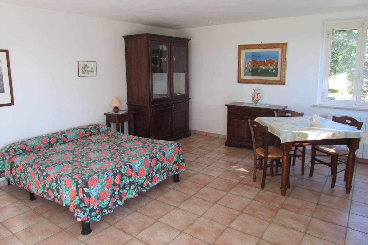 A bedroom with a bed, a wooden cabinet, a dining table with chairs, and a window.