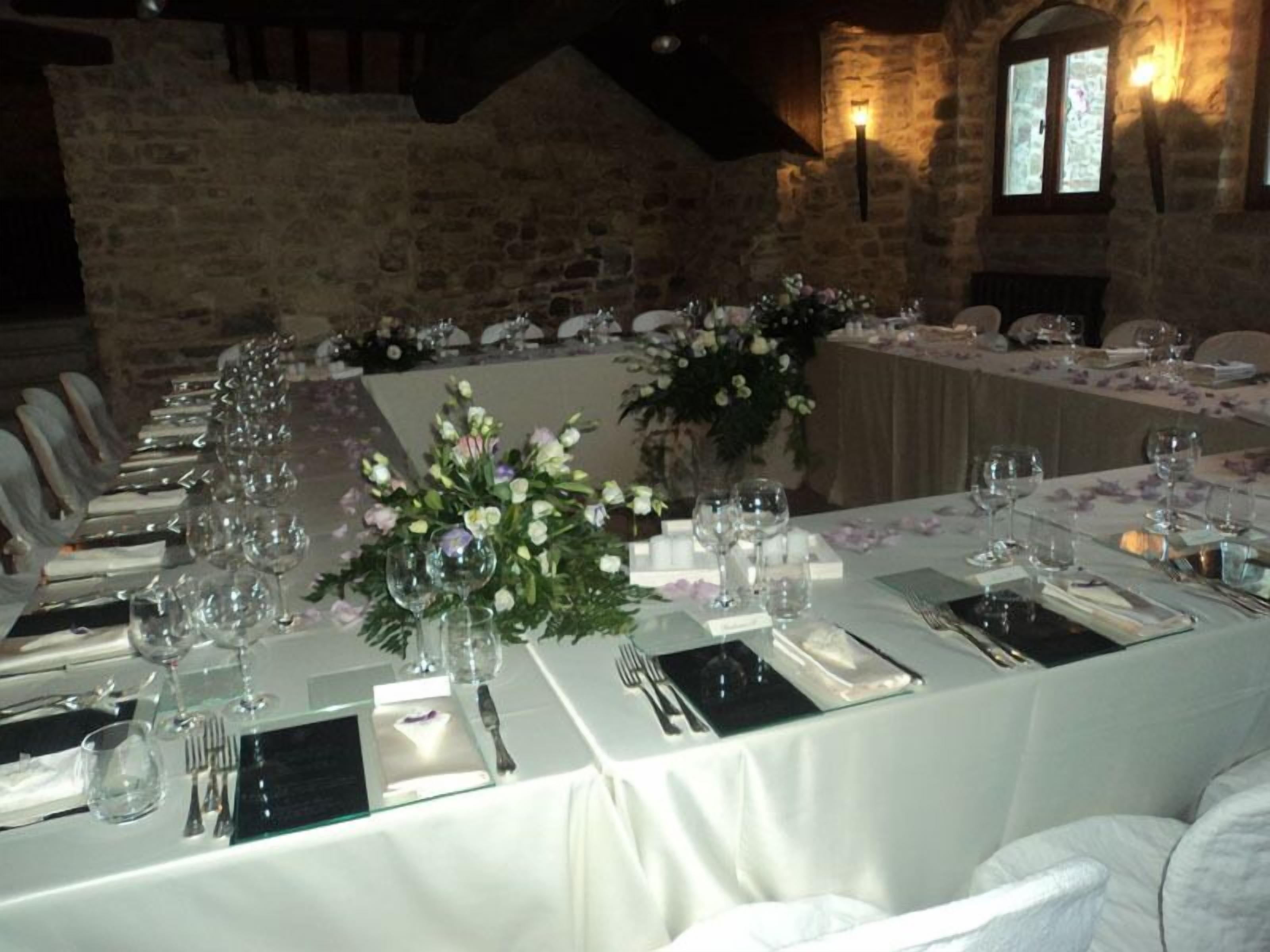 Une longue table à manger, recouverte de nappes blanches, agrémentée de verres et de compositions florales, se trouve dans une pièce aux murs de pierre, dotée d’une cheminée.