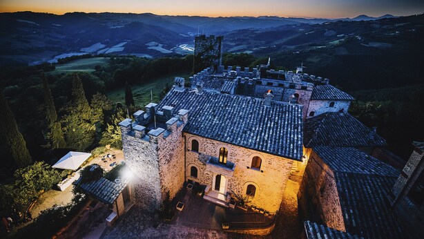 A historic stone building with a courtyard, surrounded by hills and a castle in the background. - Castello di Giomici (Valfabbrica)