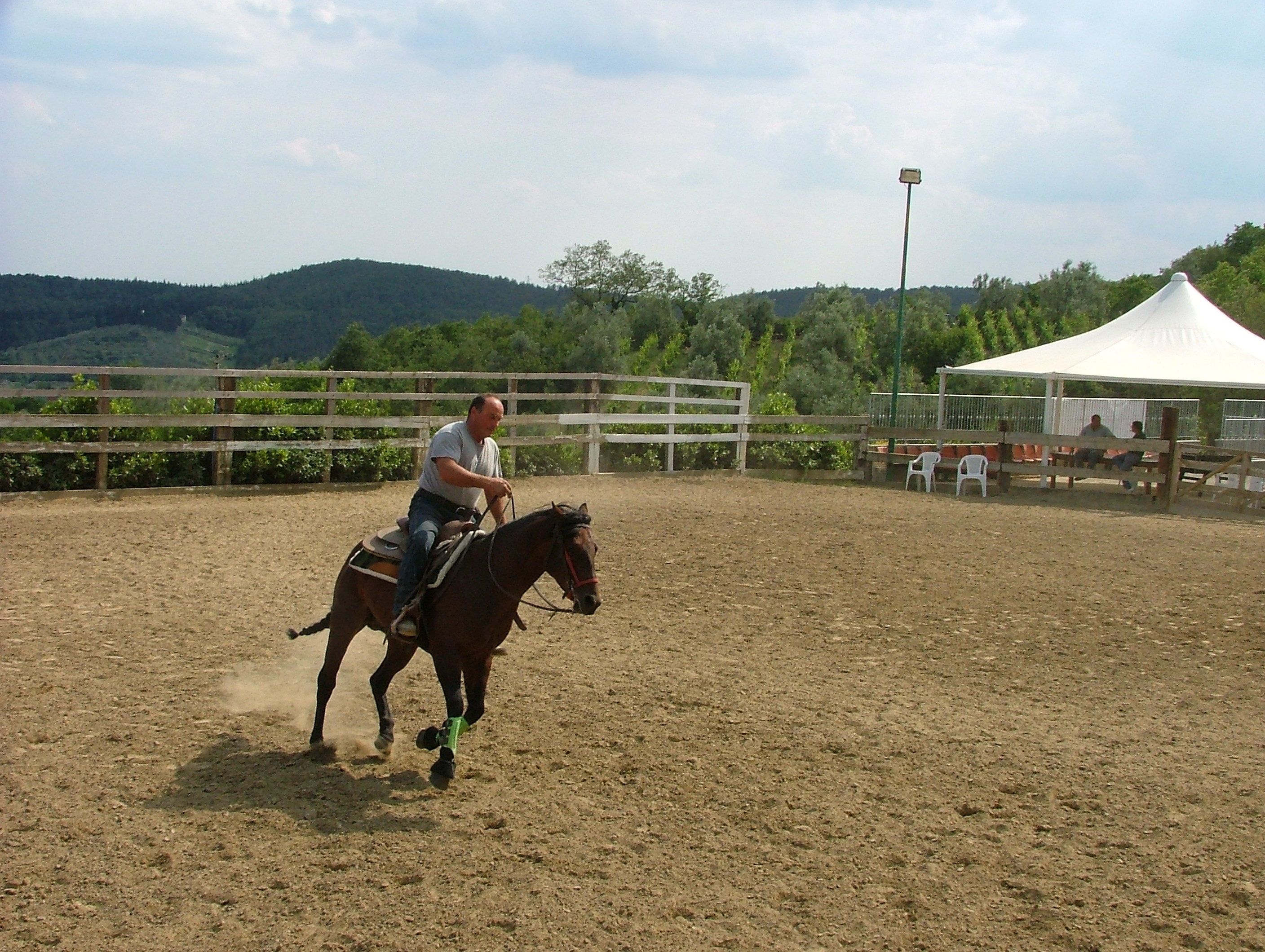 A person riding a horse in an outdoor arena with a dirt floor, surrounded by a wooden fence, and a white tent in the background.