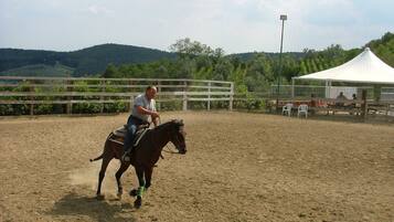 A person riding a horse in an outdoor arena with a dirt floor, surrounded by a wooden fence, and a white tent in the background.